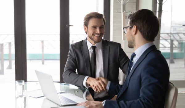 two men in suits sitting at table shaking hands and smiling