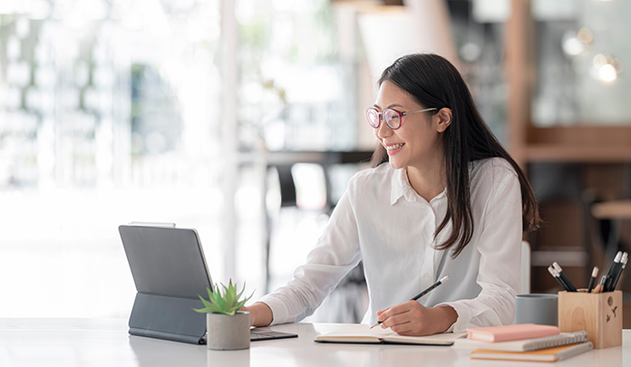 A smiling woman writes in a notebook and works on a computer